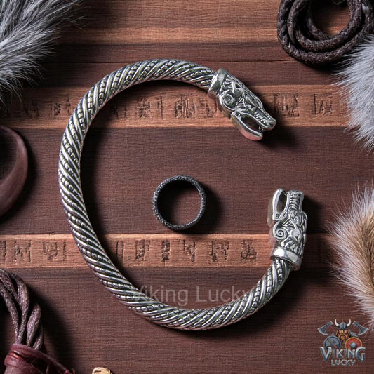 Intricate silver Viking armring with detailed dragon serpent heads on a dark wood background with runes and fur.